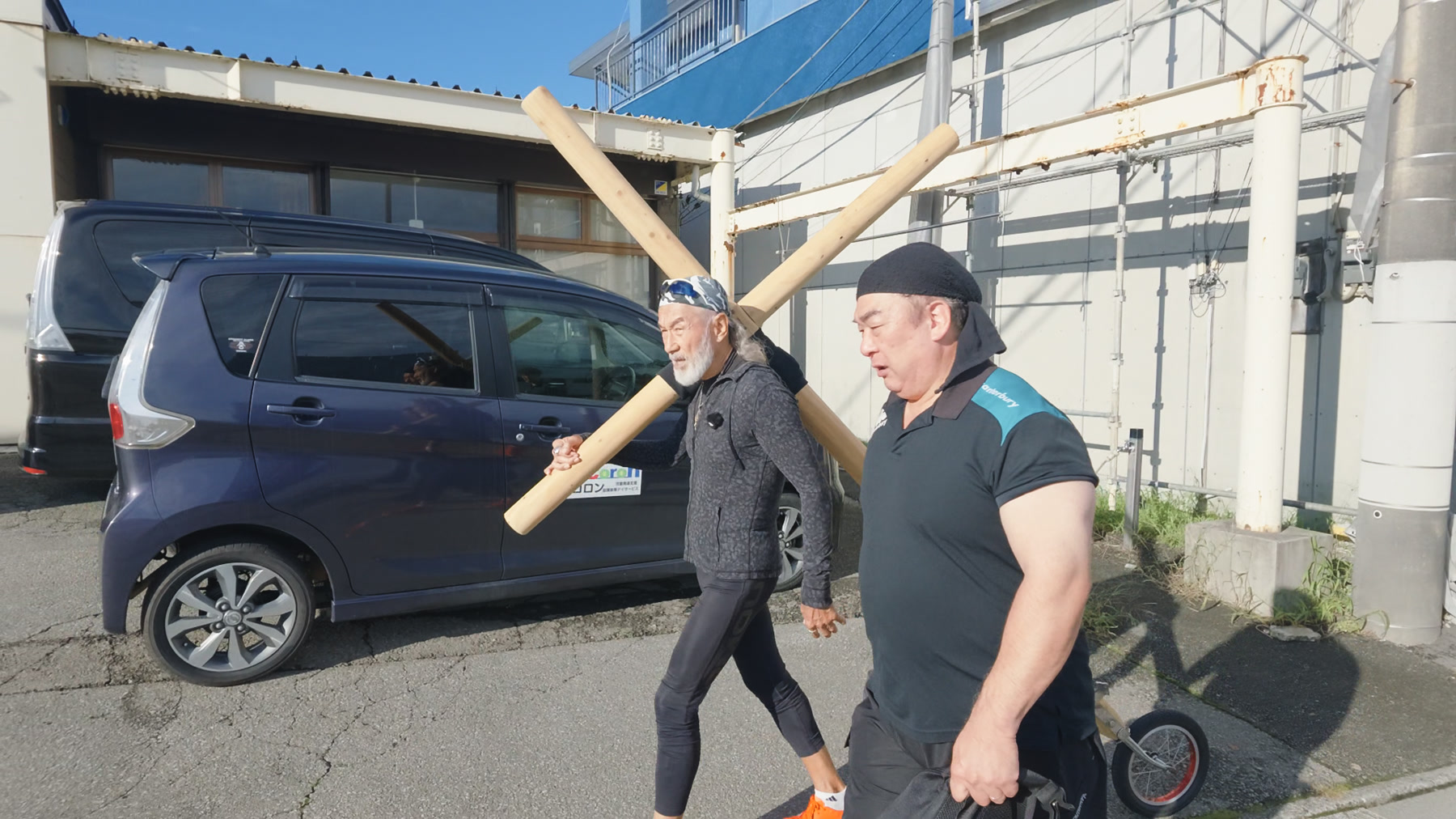 木の十字架を右肩に載せて町の道を歩くアーサー先生
